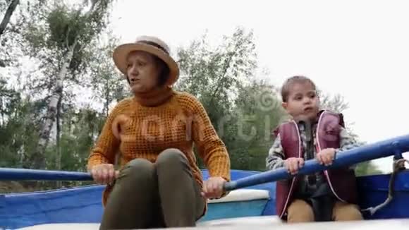 祖母漂浮在蓝色的木船上孙子在公园的湖面上在公园的船上快乐的一家人奶奶和孙子说话视频的预览图