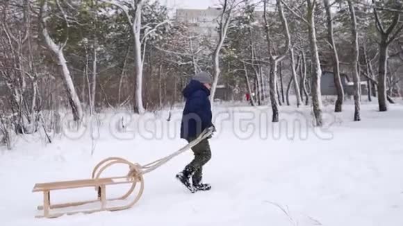 穿暖和衣服的可爱男孩在下雪的冬天拉木雪橇小男孩在公园里下雪跟踪视频的预览图