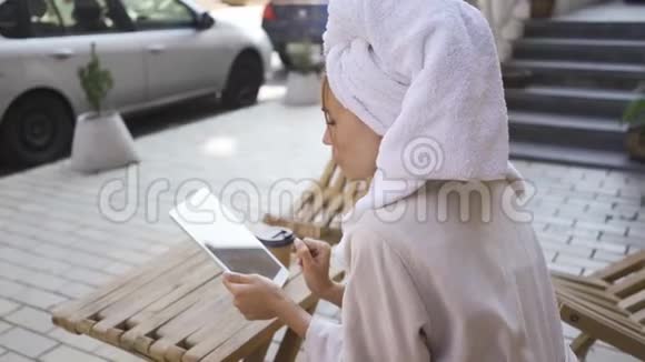 穿着浴袍头上有毛巾的可爱年轻女人的肖像坐在桌子旁后院里用平板电脑自信视频的预览图