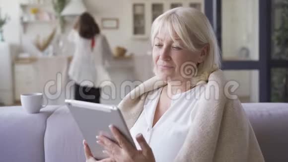 积极的成熟女人被毯子覆盖坐在沙发上看着平板电脑上的照片而她的女儿在跳舞视频的预览图