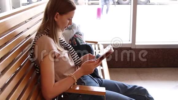年轻的金发女孩坐在长凳上等待航班在机场使用平板电脑视频的预览图
