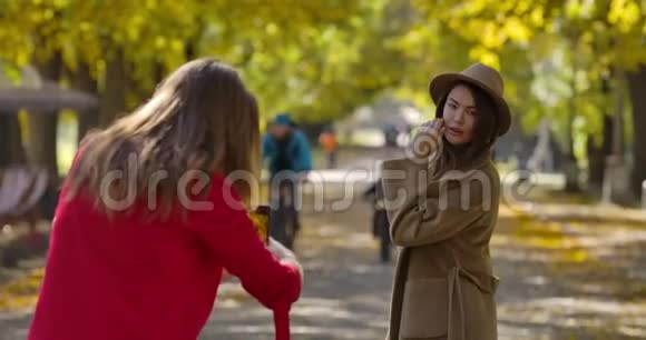 美丽的白种人女孩穿着优雅的衣服在秋天公园的背景下为她的女性朋友拍照视频的预览图