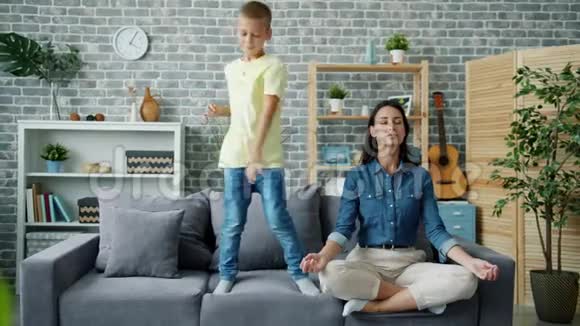 快乐的孩子在沙发上跳舞而母亲在家里坐在莲花的位置上视频的预览图