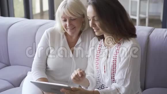 年轻的女人和她的母亲坐在沙发上在平板电脑上展示她的照片女人微笑着友好家庭视频的预览图