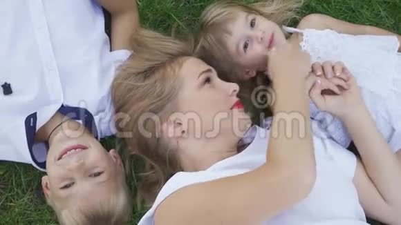 漂亮的金发女人和她的两个孩子躺在公园的草地上小男孩小女孩和成年女人视频的预览图