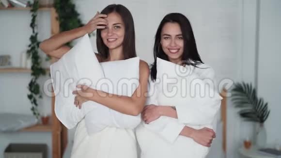 两个漂亮的女孩穿着浴袍手里拿着枕头看着相机微笑着视频的预览图