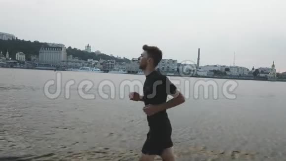 穿着黑色运动服装的性感苗条男孩沿着河滩奔跑运动员在河边跑步运动员做户外运动视频的预览图