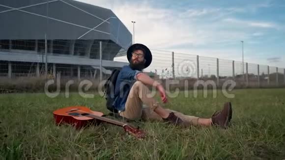 一张漂亮的胡子脸随意地放下吉他坐在草地上在大自然中休息视频的预览图