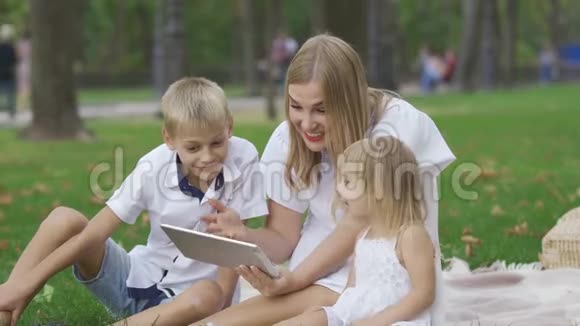 漂亮的高加索女人和她的两个孩子花时间在户外她在平板电脑上给孩子们看照片视频的预览图