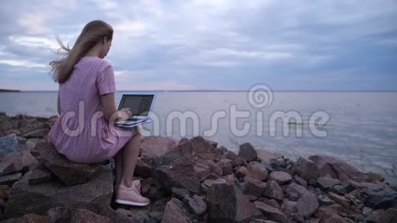 金发女郎坐在海边的石头上她正在笔记本电脑上打字寻找灵感4K慢慢视频的预览图