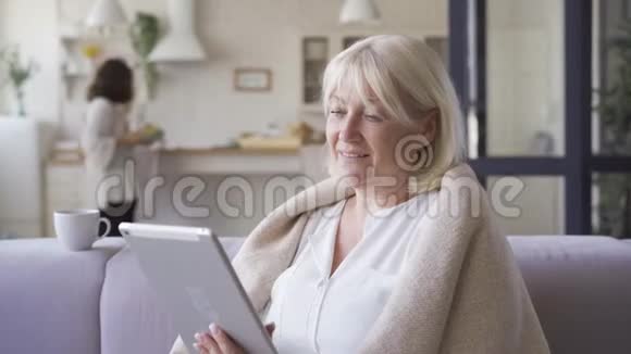 一幅成熟的女人坐在沙发上用毯子盖着的肖像看着平板电脑上的照片而她的女儿视频的预览图