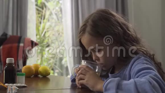 一个可爱的小女孩坐在家里的桌子旁喝着洋甘菊茶药片柠檬和橙子视频的预览图