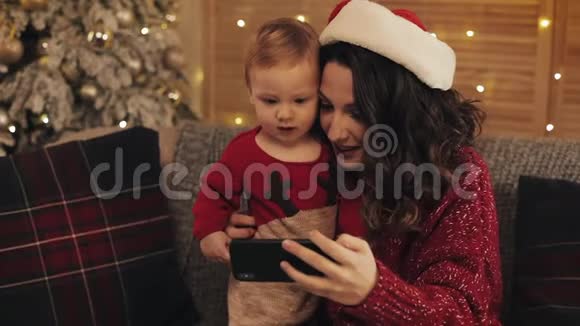 在舒适的圣诞之家美丽的母亲和可爱的小儿子微笑着坐在沙发上看着沙发视频的预览图
