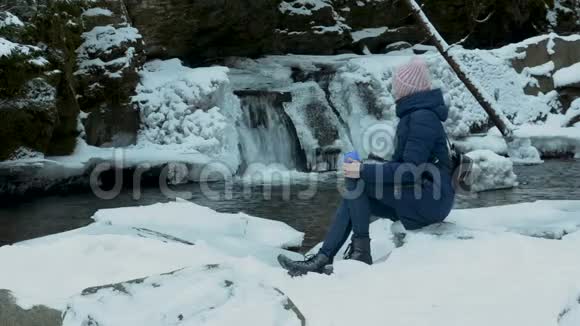 穿着蓝色外套的女孩坐在一片冰雪覆盖的针叶林中脸上挂着一个杯子背景是冰冷的瀑布和岩石冬季视频的预览图