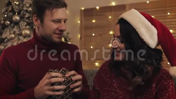 一对可爱的情侣亲吻丈夫感谢妻子在圣诞树旁的小沙发上与她在一起视频的预览图