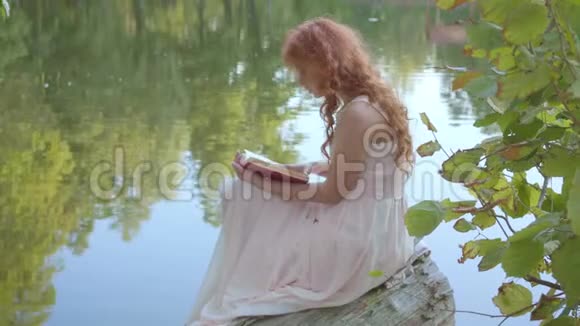 从侧面看红发迷人的白种人女孩在湖畔的深红色封面上看书视频的预览图
