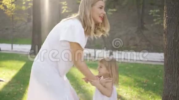 年轻的金发女人抱着她穿着白色裙子的可爱女儿旋转的肖像户外休闲幸福家庭视频的预览图