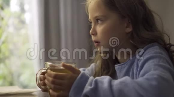 一个灰眼小女孩的特写端着一杯热茶坐在桌旁穿着蓝色毛衣的可爱孩子享受着热辣视频的预览图