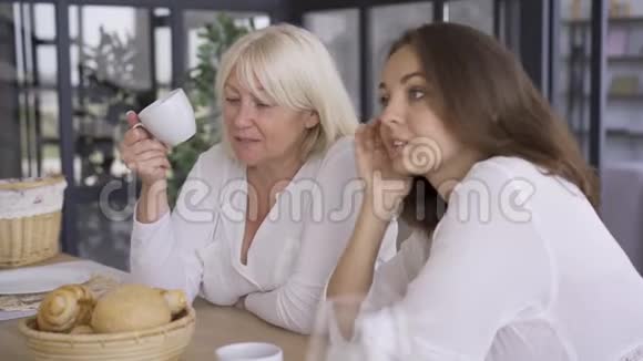 漂亮的成熟女人坐在厨房里和女儿说话喝茶或咖啡女孩和她分享视频的预览图