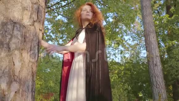 美丽的白种人女人红色的头发穿着白色的长裙黑色的长袍站在森林里看着远处休闲视频的预览图