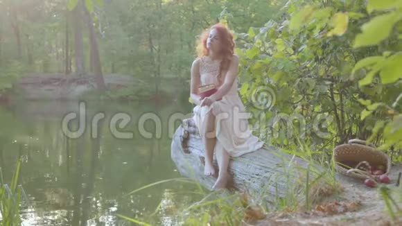 迷人的红发白种人女人穿着白色长裙坐在湖畔穿着暗红色的封面看书视频的预览图
