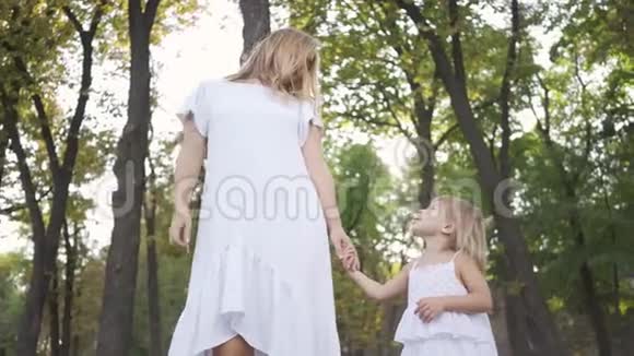 漂亮的金发女人和穿着白色连衣裙的小女孩站在公园或森林里手牵手微笑着快乐视频的预览图