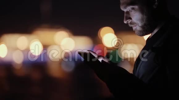 一个英俊男人的侧视微笑着点头晚上用他的平板电脑城市灯光模糊现代技术视频的预览图