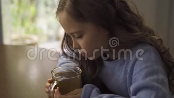 白种人的小女孩长着卷曲的黑发坐着一杯洋甘菊茶穿着蓝色衣服的可爱悲伤的孩子视频的预览图