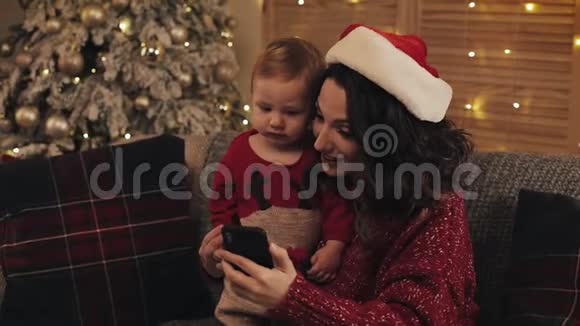 圣诞老人妈妈戴着圣诞老人帽和可爱的小儿子坐在圣诞树附近的客厅沙发上微笑快乐视频的预览图