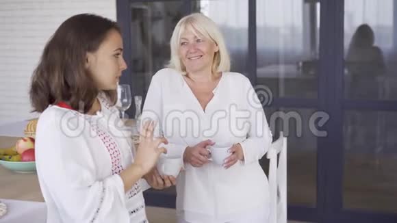 年轻的女人在喝茶或咖啡时对她母亲讲一些有趣的事情年长的女人对着镜头微笑二视频的预览图