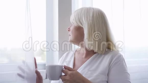 体贴成熟的女人拿着一杯茶或咖啡看着窗户妈妈在想她的孩子视频的预览图