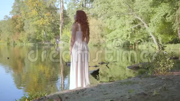 湖畔矗立着一位年轻的白种人妇女她留着一头红色的头发穿着长裙美丽的干视频的预览图