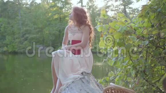 红发白种人的女人坐在河岸的原木上拿着一本深红色封面的书智能和视频的预览图