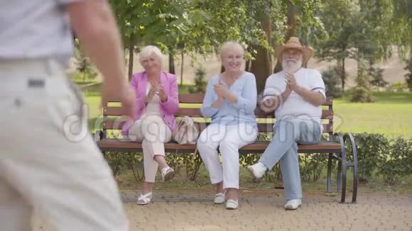三个白种人高年级学生坐在长凳上为他们欢快的男性朋友跳舞鼓掌安老公司视频的预览图