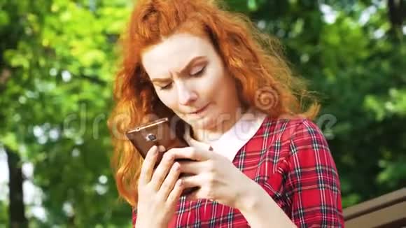 坐在公园的智能手机上有红色头发打字信息的快乐可爱女孩视频的预览图
