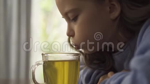 一个留着长长卷发的小白种人女孩的特写吹在一杯洋甘菊茶上品尝它可爱的悲伤视频的预览图