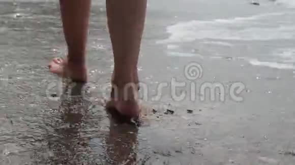 在一个年轻女孩美丽的脚后拍摄她沿着沙质的海岸沿着大海的泡沫小浪走视频的预览图