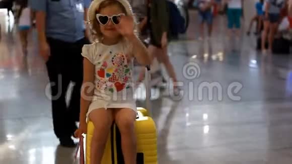 小女孩游客坐在火车站的一个黄色的大行李箱上挥手告别孩子正在等待视频的预览图