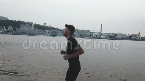 穿着黑色运动服装的年轻运动男孩沿着河滩奔跑活泼苗条的男人在河边跑步户外运动的运动员视频的预览图