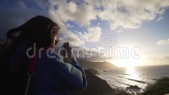 年轻的时髦女人在电话里拍摄日落时海岸令人景色吹毛求疵加那利视频的预览图
