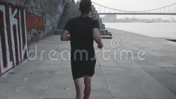 职业运动员在码头跑步的后视图活泼苗条的男人沿着河跑运动健康的小男孩在长廊上奔跑视频的预览图