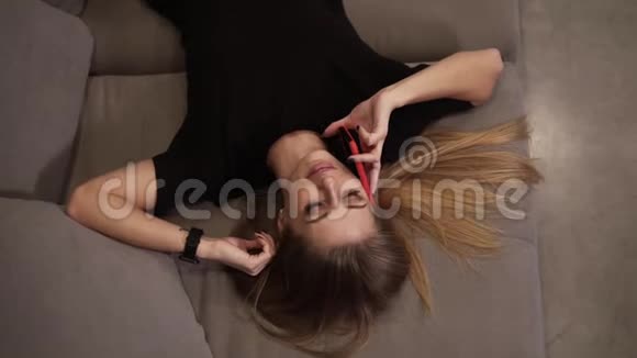 漂亮性感的女孩带着手机躺在家里的沙发上穿着黑色衣服的金发放松的女孩在手机旁讲话视频的预览图
