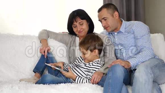 母亲父亲和孩子坐在沙发上用平板电脑看电影玩或使用互联网快乐视频的预览图
