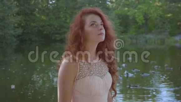 美丽的白种人女人红色的头发穿着米色的连衣裙站在湖边抬头看着周围红头发视频的预览图
