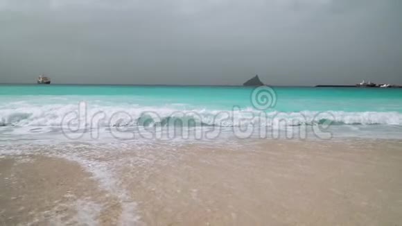 蓝绿色的海浪缓慢地向白色的沙滩滚动背景是巨大的岩石视频的预览图