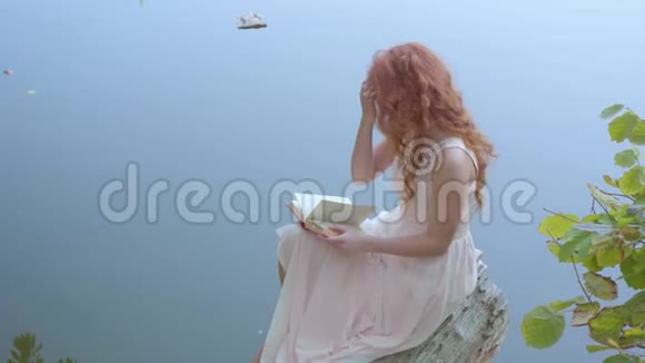 侧面看一个年轻迷人的女孩留着长长的红色头发在蓝湖的背景下阅读诗歌可爱的白种人视频的预览图