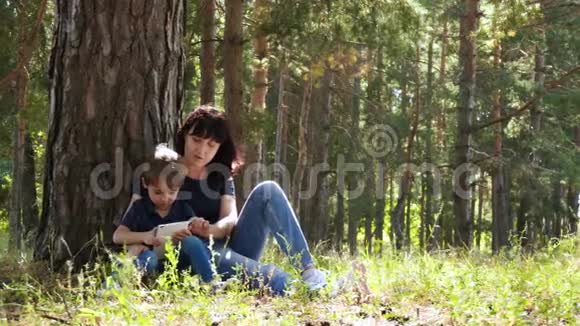 一位年轻的母亲和她的儿子正在公园里放松坐在一棵树旁用一个小玩意当代科技视频的预览图