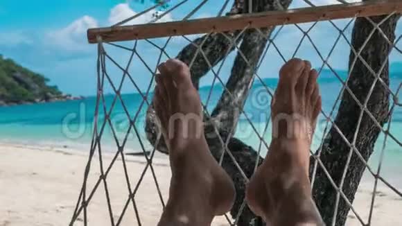 在白沙滩和蔚蓝的海洋前光着脚在吊床上摇摆视频的预览图