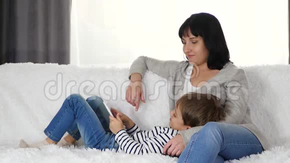 一位母亲抱着儿子坐在房间里的沙发上小男孩在玩平板电脑现代幸福的概念视频的预览图