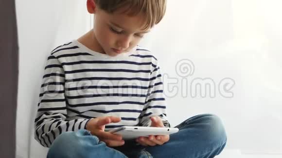 一个小男孩看着平板电脑的屏幕坐在窗边这孩子用手指上下抚摸视频的预览图